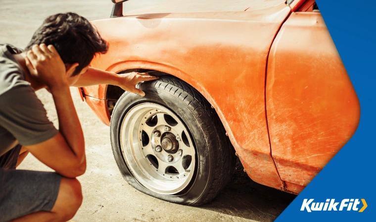 Person crouches beside their car, inspecting a flat tyre.